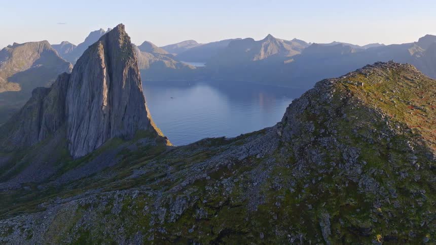 A stunning panoramic view from a rocky mountain summit reveals a majestic peak and a serene fjord below under a clear sky