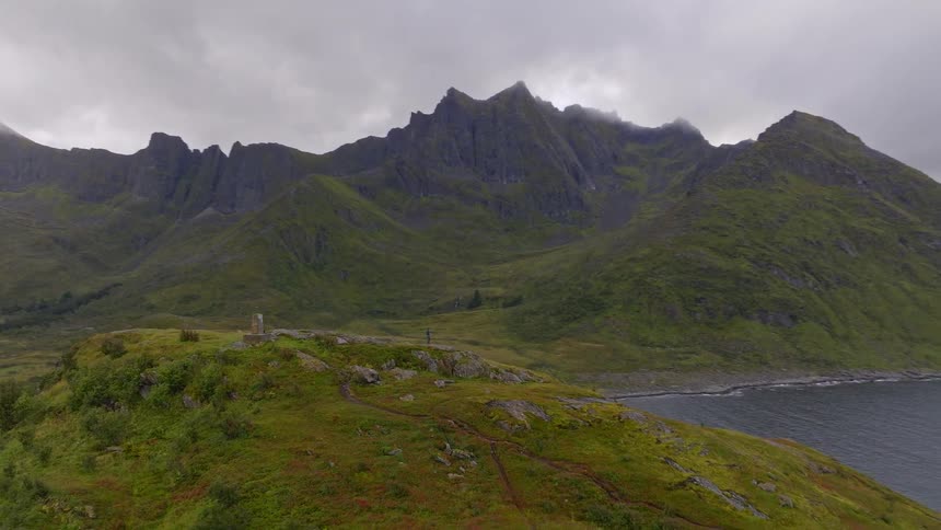A slow aerial pullback reveals a solitary hiker dwarfed by the immense and dramatic green mountains along a rugged coastline