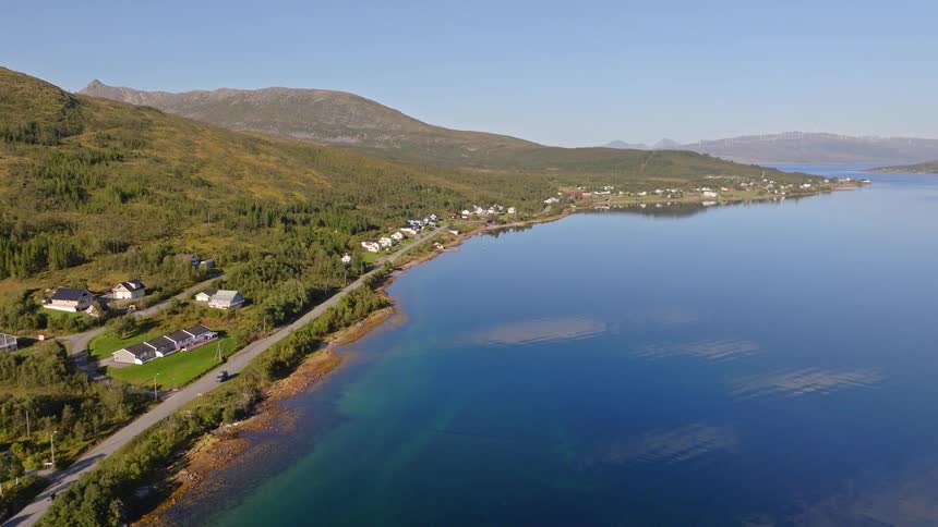 An aerial shot slowly pulls back from a quaint village situated on the picturesque coastline of a Norwegian fjord under a clear blue sky