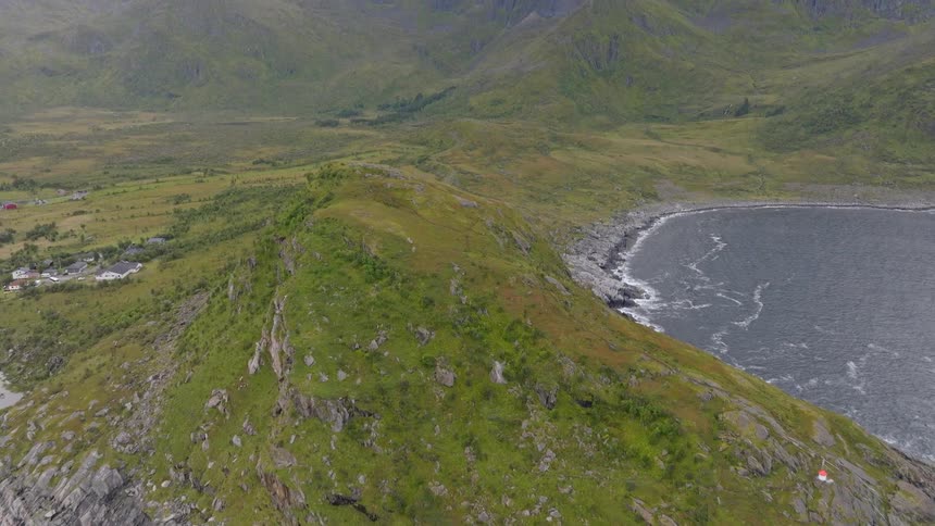 Aerial drone rises over a rocky, green coastal peninsula in Norway to reveal a stunning, epic mountain range