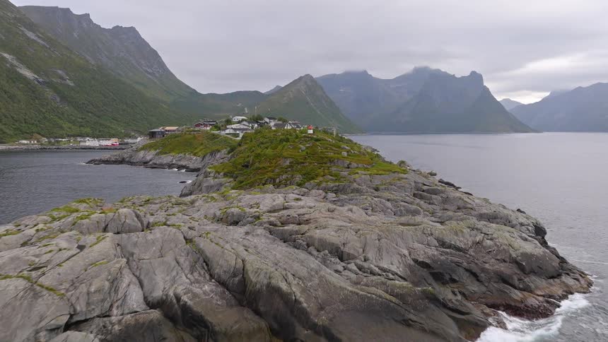 Aerial footage pulls away from a man at a small lighthouse on a rocky peninsula to reveal the vast, mountainous landscape of a Norwegian fjord