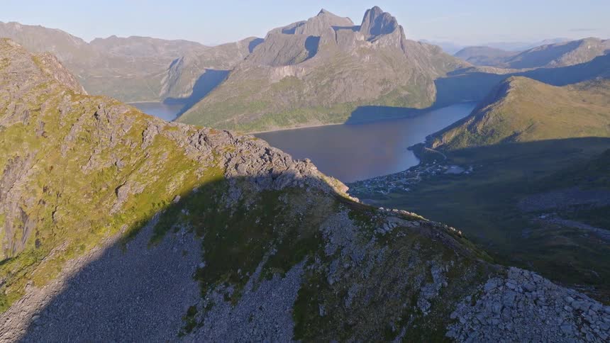 Aerial tilt shot reveals a stunning fjord and coastal village surrounded by vast, rugged mountains at sunrise