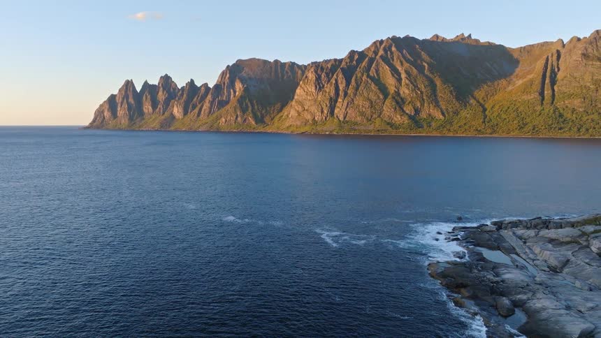 A stunning aerial view pulls back over the Norwegian Sea to reveal the jagged mountain peaks of Tungeneset at golden hour