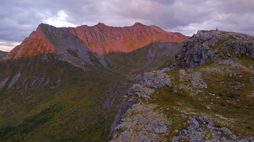 Epic aerial shot over a rocky ridge towards a breathtaking mountain range glowing at sunset