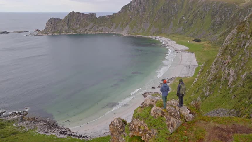 Two hikers stand on a dramatic, grassy cliff edge, admiring the magnificent view of a secluded sandy beach and mountainous coastline