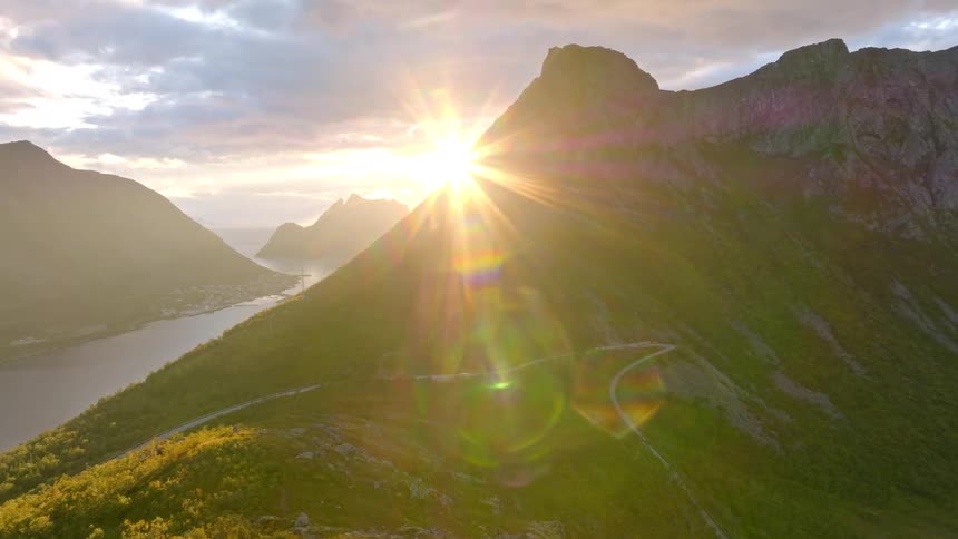 Awe-inspiring aerial view reveals a brilliant sunburst emerging from behind a majestic mountain, casting golden light over a serene Norwegian fjord and landscape