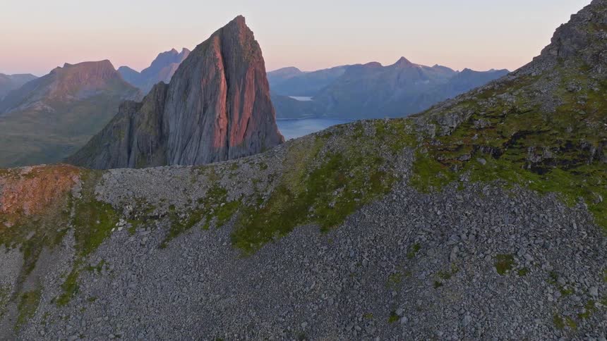 An aerial drone shot flies over a rugged mountain ridge towards a majestic peak illuminated by the warm glow of sunset, revealing a deep blue fjord below