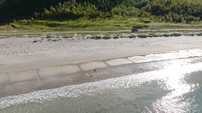 Epic aerial reveal of a lone person walking along a pristine beach, dwarfed by a majestic mountain landscape