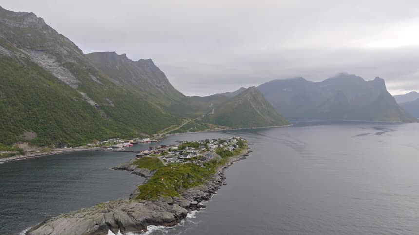 Aerial drone pulls back revealing a remote village on a rocky peninsula surrounded by a stunning Norwegian fjord and mountains