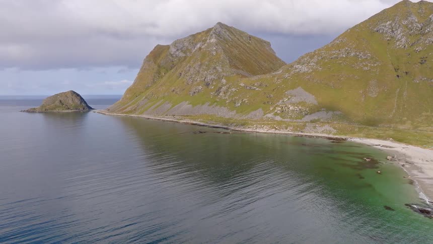 Majestic green mountains rise from a tranquil sea next to a pristine sandy beach under a cloudy sky