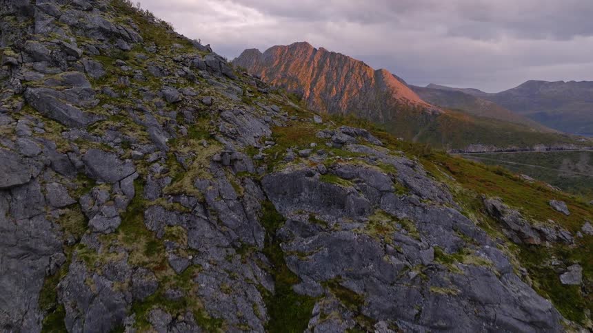 An aerial view ascends a rocky mountainside to reveal a spectacular sunlit mountain range and fjord at dusk