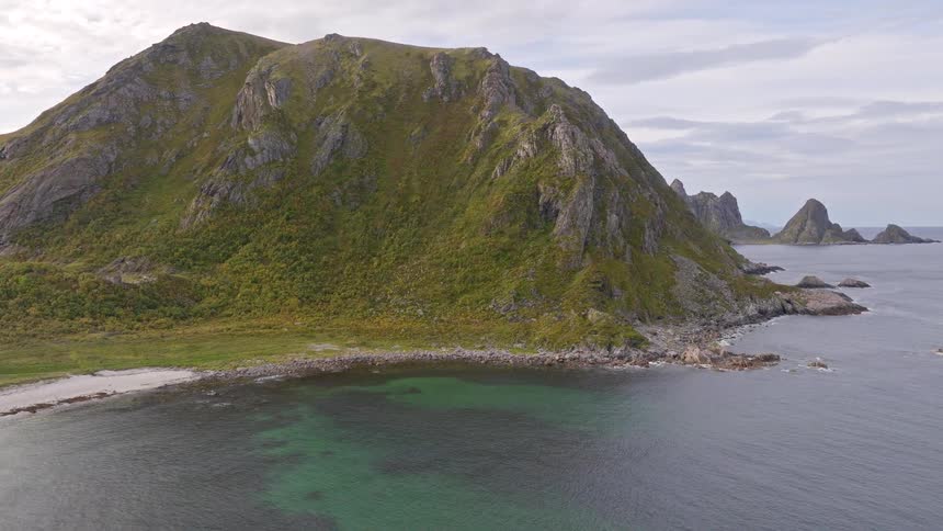 A spectacular aerial view reveals a lush green mountain towering over a secluded sandy beach and the clear waters of the Norwegian Sea