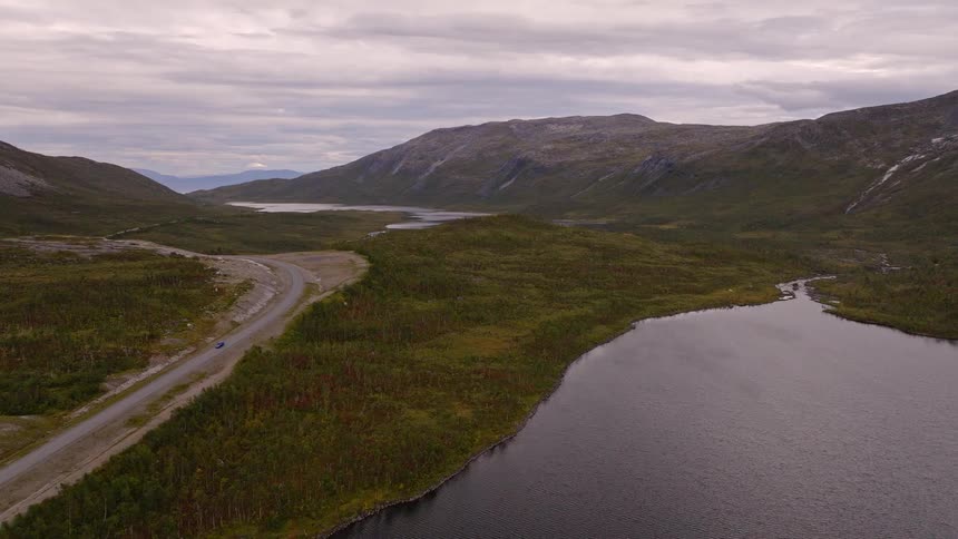 An aerial view ascends over a vast and moody mountain valley, revealing a serene lake and a winding road through the remote wilderness