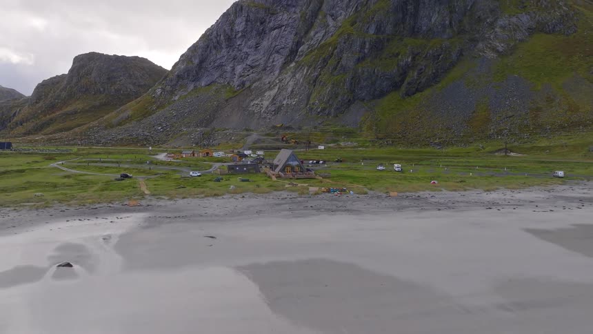 Aerial view pulls back from a coastal campground and lodge, revealing a vast sandy beach and dramatic mountains in the background