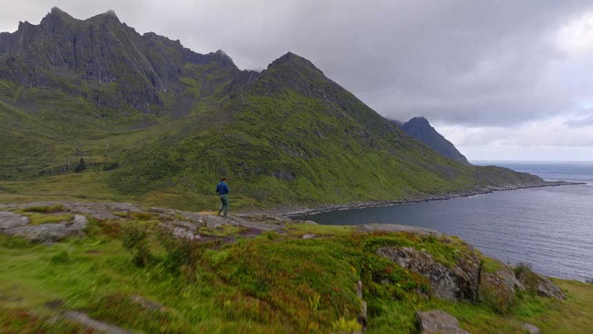 A drone flies over a lone hiker standing atop a lush green cliff, revealing a stunning view of a dramatic coastline and towering mountains