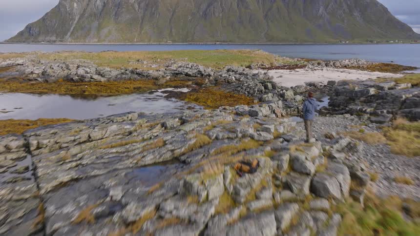 Aerial view flies over a rocky shoreline covered in yellow seaweed, revealing a lone hiker admiring the majestic mountain view across the water