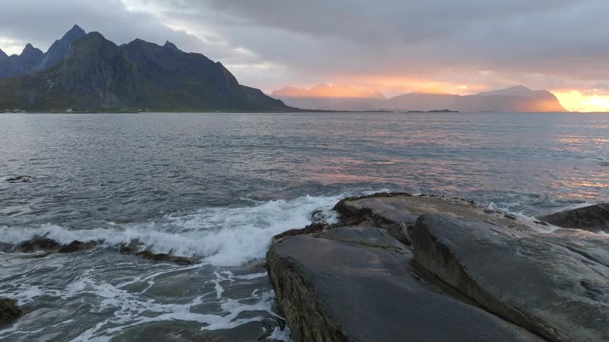 A slow-motion aerial shot moves over the rocky coastline of a fjord as waves crash and the sun sets behind dramatic mountains