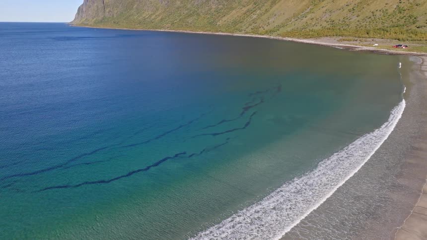 An aerial view pulls back from the tranquil turquoise waters and sandy shores to reveal a breathtaking mountain range along a pristine coastline