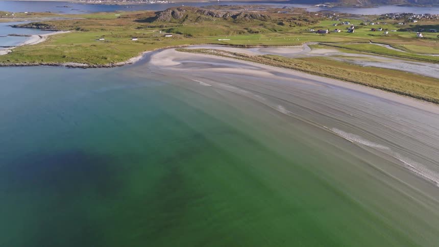 A breathtaking aerial view tilts up from the clear turquoise water to reveal a stunning sandy beach and dramatic mountain landscape in Norway