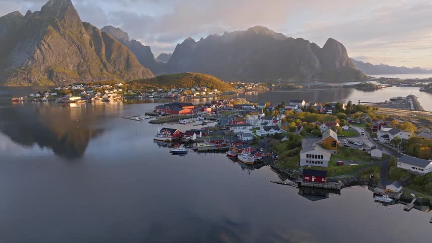 A stunning aerial view pans across a tranquil fishing village in the Lofoten Islands, Norway, as sunrise illuminates the dramatic mountains and their reflections in the calm water