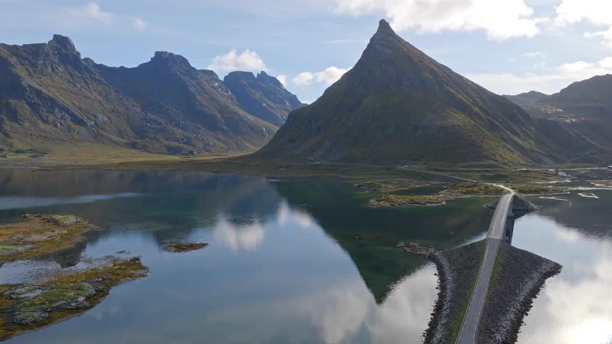 Stunning aerial view of a majestic mountain peak and its perfect reflection in a tranquil fjord, with a scenic road and bridge crossing the water in Norway