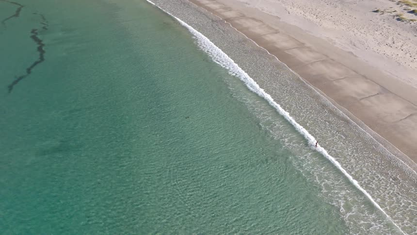 An aerial view captures a lone person wading in the clear turquoise water of a remote beach before the camera ascends to a top-down perspective of the vast landscape
