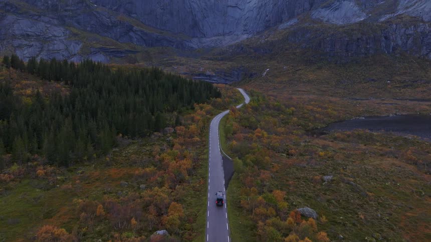 An aerial shot follows a camper van driving along a remote, winding road through an autumn valley, revealing majestic, cloud-covered mountains in the distance