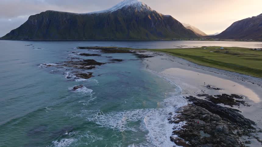 A breathtaking aerial view tilts up to reveal a snow-capped mountain overlooking a beautiful beach with turquoise waters in Lofoten, Norway