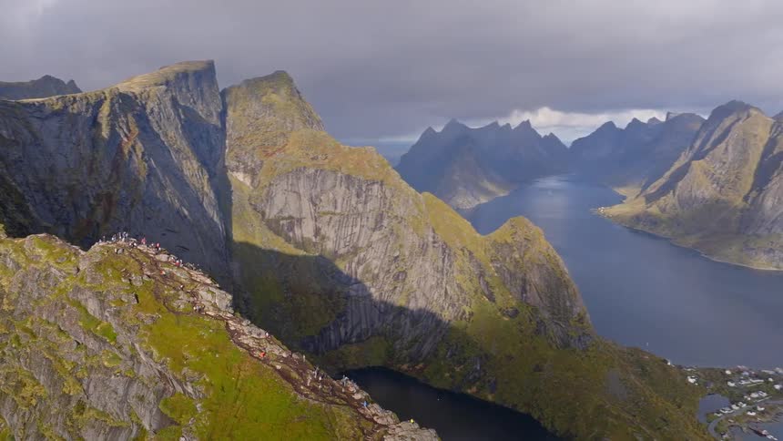A breathtaking aerial shot pulls back from hikers on the Reinebringen mountain ridge, revealing the magnificent Lofoten Islands landscape in Norway