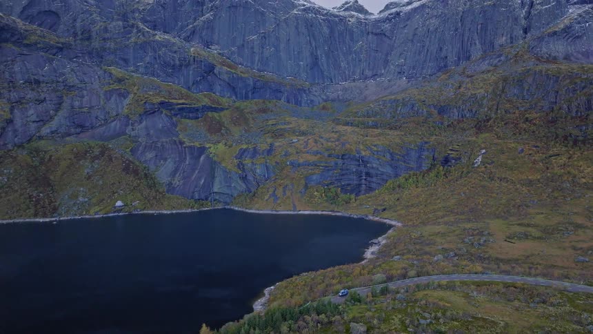 A scenic aerial view follows cars on a winding road alongside a lake, tilting up to reveal the stunning, snow-dusted mountains of Lofoten, Norway