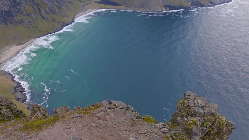 Aerial view pans up from a lone hiker on a cliff edge to reveal an epic landscape of mountains and a remote beach in Norway