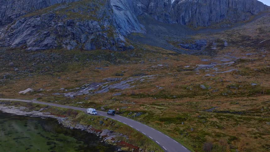 An aerial shot follows two campervans driving on a scenic road alongside a tranquil lake, revealing the majestic, snow-dusted mountains of Norway