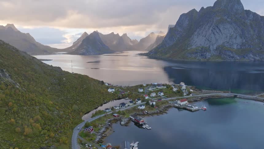 Aerial footage rises over a remote Norwegian fishing village nestled in a fjord surrounded by majestic mountains under a dramatic cloudy sky