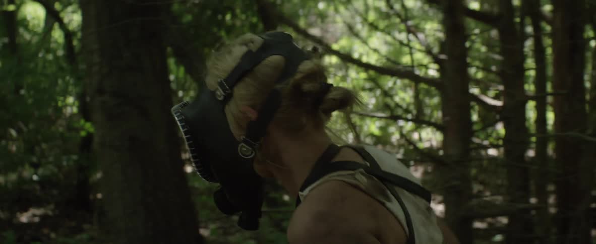 A woman wearing a type of gas mask walks in the dense forest