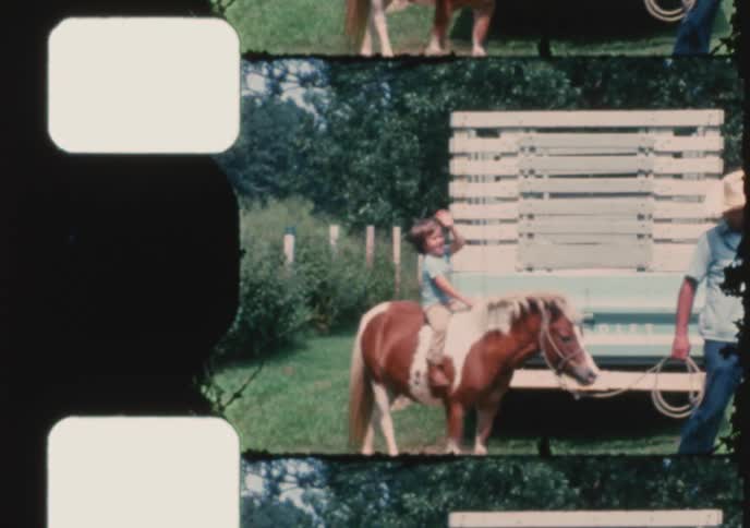 A father in a cowboy hat leads his smiling young son on a pony ride through the yard in this vintage home movie