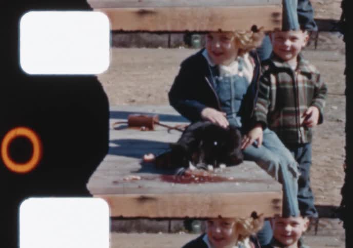 A vintage 8mm home movie shows a young girl and her brother petting and feeding their black and white cat outdoors on a sunny day