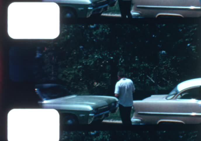 A vintage home movie from the 1960s captures a child's first pony ride, held steady by a man with classic cars parked nearby