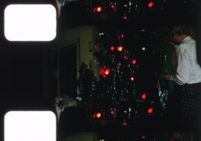 Vintage 8mm film captures a woman meticulously decorating a Christmas tree with colorful lights and silver tinsel in a cozy home