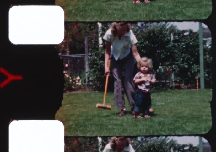 A mother teaches her toddler with curly blonde hair how to play croquet in their sunny backyard in this vintage home movie clip
