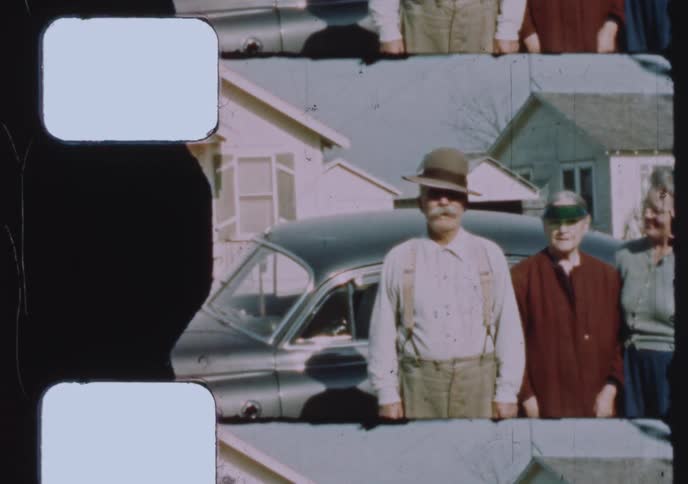 A vintage 16mm film scan shows a group of elderly friends or family posing together in front of a classic car in a residential neighborhood
