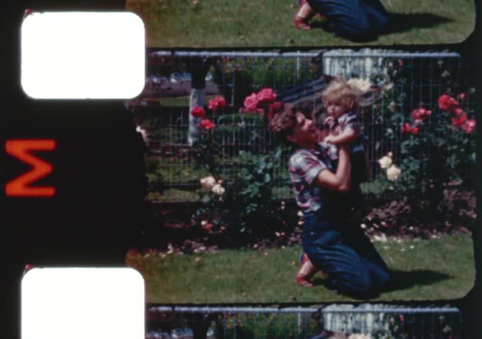 Vintage 16mm film shows a mother lovingly holding her toddler in a sunny garden full of roses