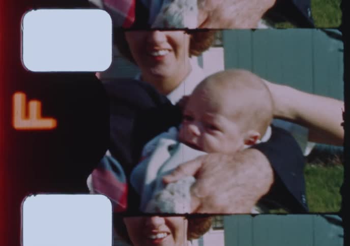 A film scan of a vintage home movie shows a proud grandfather holding a newborn baby surrounded by his smiling family