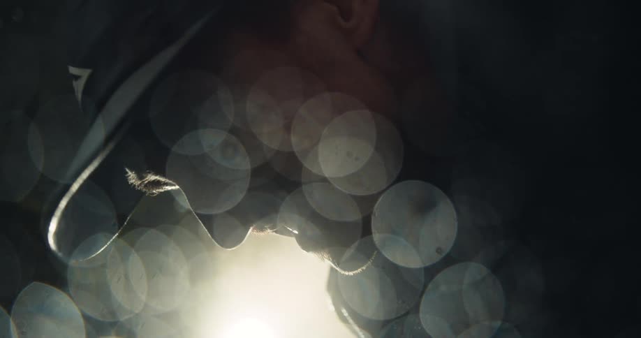 A close-up, backlit profile of a somber man in a cap, his face cast in shadow amidst atmospheric bokeh