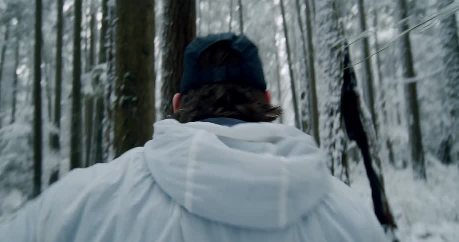 A man wearing a white jacket and baseball cap runs through a dense, snow-covered forest