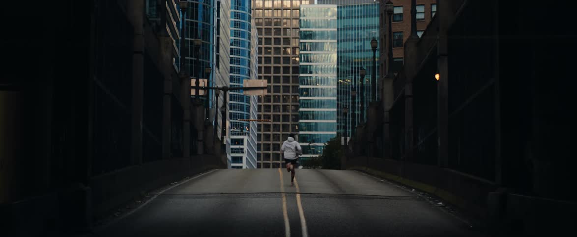 A lone runner emerges from the shadows and jogs up an empty city street towards towering skyscrapers