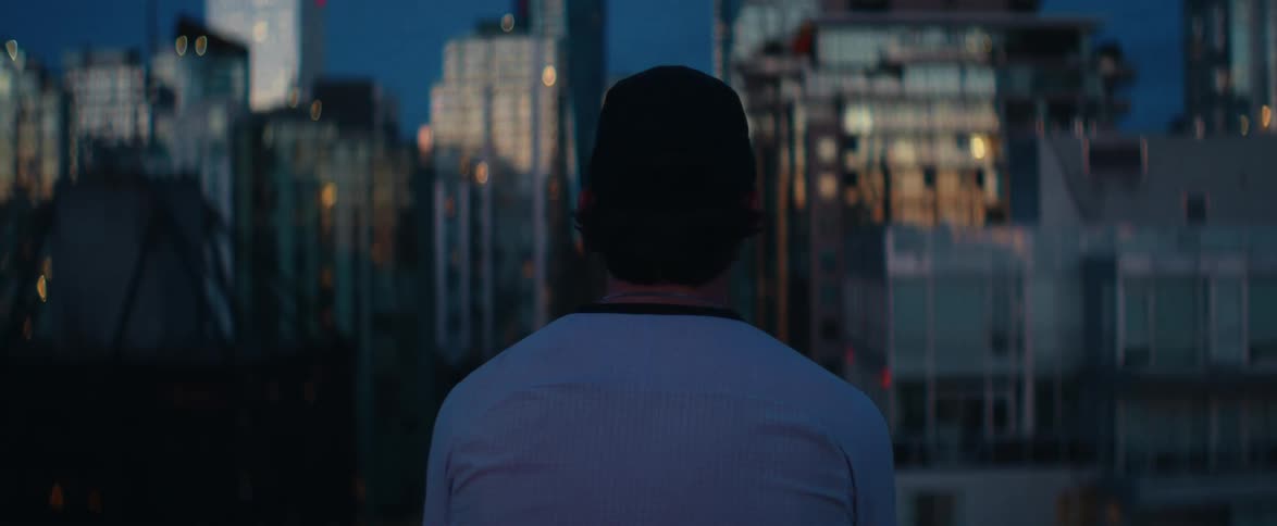 From behind, a young man in a backwards cap looks out over a modern city skyline at twilight as the camera slowly pulls focus to the distant buildings