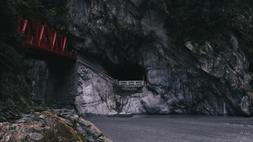 A striking red bridge stands beside a dark, flowing river leading to a mysterious cave entrance carved into a massive rock canyon