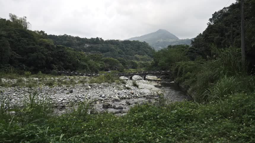 An old stone arch bridge spans a rocky river in a lush, green mountain valley on an overcast day
