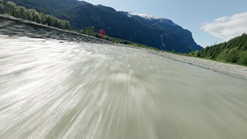 An FPV drone speeds low over a fast-flowing glacial river through a rugged mountain valley on a bright summer day