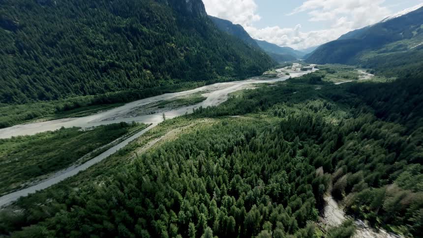 A majestic drone flight reveals a sprawling river valley carved between lush, forest-covered mountains on a sunny day